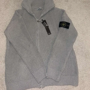 Grå stickad kofta från Stone Island - Säljer en grå stickad kofta från Stone Island med dragkedja och hög krage. Klassisk patch på ärmen och grovstickad struktur ger en clean och stilren vibe. Perfekt för lager-på-lager och snygg till jeans eller cargobyxor.