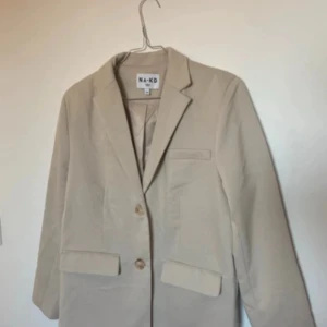 Beige kavaj från NA-KD, stl 36 - Stilren beige kavaj från NA-KD i klassisk modell med två knappar framtill och fickdetaljer. Perfekt för en clean och trendig look. Tillverkad i ett mjukt material som känns bekvämt hela dagen.