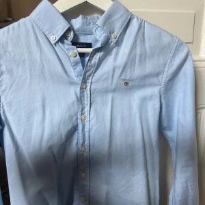 Ljusblå skjorta från Gant - Klassisk ljusblå skjorta från Gant med broderad logga på bröstet. Skjortan har button down-krage, knappar framtill och långa ärmar. Perfekt för en clean och stilren look.