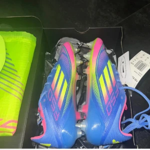 Adidas F50 färgstarka fotbollsskor - Snygga Adidas F50 fotbollsskor i blått, rosa och neongult. Skorna har klassiska tre ränder på sidan, snörning och en lätt ovandel i syntetmaterial. Perfekta för dig som vill sticka ut på planen med grymma färger och modern design.