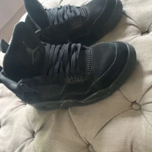 Svarta Nike Air Jordan 4 sneakers - Säljer ett par äkta black cats Nike Air Jordan 4 sneakers med klassisk siluett och coola detaljer. Skorna har snörning, meshpaneler och är tillverkade i mocka och syntet. Perfekta för dig som vill ha en ikonisk och stilren look. 