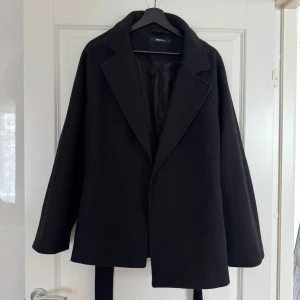 Svart kappa från Gina Tricot - Stilren svart kappa från Gina Tricot med klassisk krage och bälte i midjan. Jackan har en rak passform och är helfodrad, perfekt för kyliga dagar. Enkel och snygg design som funkar till många outfits.