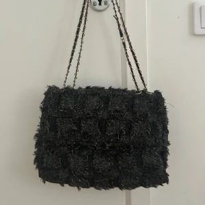 Grå axelväska med kedjerem - Säljer en svart axelväska med coolt rutigt och fransigt tyg. Väskan har en kedjerem i metall och läder, vilket ger en edgy vibe. Perfekt för dig som vill sticka ut med en unik accessoar. Rymmer mobil, plånbok och lite till.