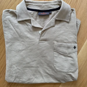 Beige t-shirt med krage från Morris - Snygg beige t-shirt från Morris med klassisk krage och knappdetalj framtill. Bröstficka med broderad svart logga ger en clean look. Tillverkad i mjuk bomull, perfekt för en chill och stilren vibe.