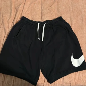 Svarta Nike shorts - Svarta Nike shorts med vit snörning i midjan och stor vit swoosh-logga på ena benet. Klassisk loose fit med elastisk midja och fickor på sidorna samt en bakficka. Perfekta för träning eller chill dagar. Materialet är mjuk.