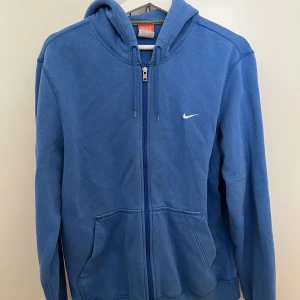 Blå Nike hoodie med dragkedja - Blå hoodie från Nike med vit logga på bröstet. Tröjan har huva med snörning, hel dragkedja och två stora fickor framtill. Perfekt för chill dagar eller när du vill ha en sportig look.
