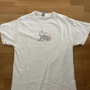 Vit t-shirt Christian Livin tryck - Vit t-shirt med rund hals och korta ärmar. På bröstet finns ett litet tryck med texten 'Christian Livin' och motiv i pastellfärger. Skön och enkel modell i mjuk bomull, perfekt för en chill look.