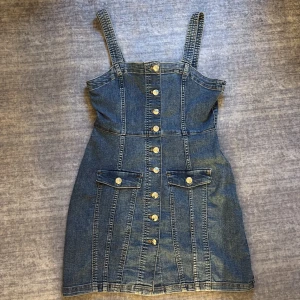 Jeansklänning med knappar från H&M - Snygg jeansklänning från H&M i klassisk blå denim. Kort modell med breda axelband, knappar hela vägen framtill och två fickor med lock. Perfekt för dig som gillar en avslappnad men trendig stil. ALDRIG ANVÄND!