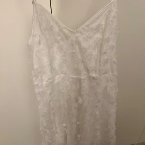 Vit klänning med blommor  - Superfin vit kortklänning med tunna axelband och v-ringning. Klänningen är täckt av spets och 3D-blommor, vilket ger en romantisk och unik look. Perfekt för dig som vill sticka ut med en elegant och detaljrik stil🤍 Storlek S