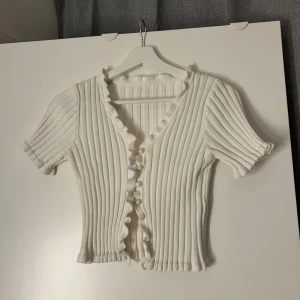 Vit ribbad blus med volang och pärlknappar - Supersöt vit ribbad blus med korta ärmar, v-ringning och volangkant längs knäppningen. Blusen har pärlknappar framtill och är croppad i modellen. Perfekt för dig som gillar romantiska detaljer och en feminin look.💞