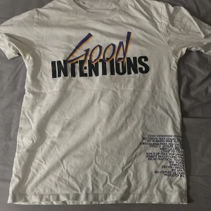 Vit Good Intentions t-shirt VLONE - Behöver bara strykas! Vit t-shirt från VLONE med stor 'Good Intentions'-text framtill i svart, lila och gult. På ryggen finns ett stort lila V med två duvor i rosa och lila. Tryckt låtlista nere till vänster både fram och bak. Klassisk passform och rund hals. Perfekt statement-plagg för streetwear.