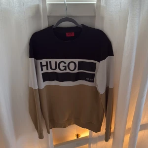 Beige och svart sweatshirt från Hugo - Snygg sweatshirt från Hugo med breda blockränder i svart, vitt och beige. Stor logga framtill och text på ryggen. Klassisk rund hals och långa ärmar. Perfekt för dig som gillar streetwear och vill sticka ut med en stilren look.