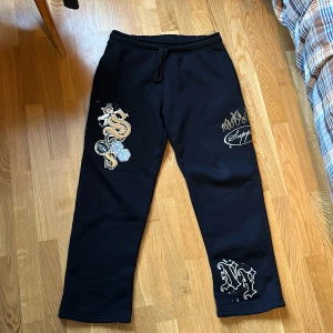 Svarta mjukisbyxor Supply & Demand S - Svarta jättesköna mjukisbyxor från Supply & Demand med streetinspirerade tryck som dollartecken, tärningar, flames och NY-text. Byxorna har dragsko i midjan och raka ben. Perfekta för en chill och trendig look. Insidan är väldigt sköna och mjuk material.