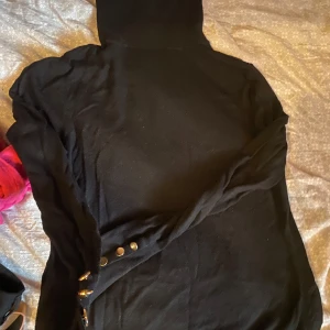 Svart hoodie med knappar på ärmen - Svart hoodie med lång ärm och coola metallknappar längs ena ärmen. Tröjan har huva och är i ett mjukt material som känns skönt mot huden. Perfekt för dig som gillar en enkel men ändå unik stil.