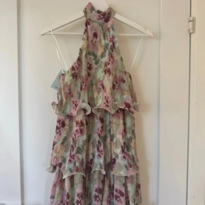 Blommig volangklänning med halterneck - Supersöt blommig klänning från Gina tricot med volanger i lager, halterneck och öppen rygg. Färgerna går i ljusrosa, vinrött och grönt på en ljus bas. Perfekt för sommarens alla tillfällen när du vill sticka ut lite extra. Nypris 600
