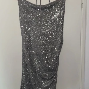 Glittrig silver kortklänning - Supersnygg kort klänning i silver med massor av paljetter som glittrar. Klänningen har halterneck och en tight passform som framhäver figuren. Perfekt för dig som vill sticka ut och glänsa på festen eller nyår.