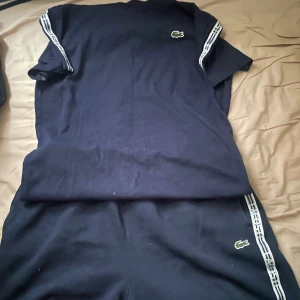 Mörkblå Lacoste t-shirt och Shorts - Snygg mörkblå set från Lacoste med klassisk krokodillogga på bröstet. T-shirten har korta ärmar och vita ränder med Lacoste-tryck längs axlarna. Och samma med shortsen så som på bild  Den är för liten på mig och ligger bara där i garderoben