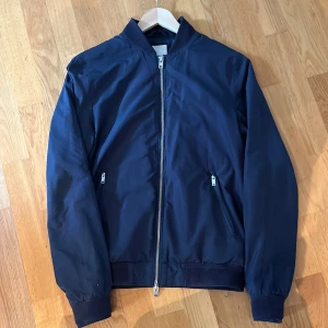 Jacka från J.Lindeberg - Snygg marinblå jacka från J.Lindeberg. Jackan har dragkedja framtill och två fickor med dragkedja. Den är tunt material så perfekt för sena sommarkvällar, ny pris cirka 1600, utmärkt skick (9/10) pris kan diskuteras