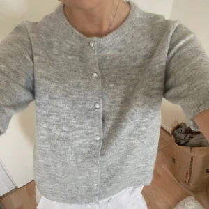 Ljusgrå stickad kofta med pärlknappar - Superfin ljusgrå stickad kofta med korta ärmar och pärlknappar framtill. Koftan har en rund halsringning och är i en mjuk, mysig stickad kvalitet. Perfekt att slänga över en topp.
