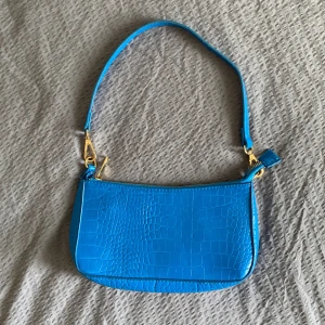 Blå axelväska i croco-imitation - Snygg blå axelväska med croco-mönstrad yta och guldfärgade metalldetaljer. Väskan har en avtagbar axelrem och stängs med dragkedja. Perfekt storlek för det viktigaste och ger en cool färgklick till din outfit.