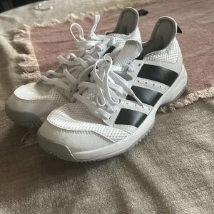Vita Adidas sneakers med svarta ränder - Snygga vita Adidas sneakers med klassiska svarta ränder på sidorna. Ovandelen är i mesh och syntet för bra andningsförmåga och komfort. Skorna har snörning och en slitstark, mönstrad sula som ger bra grepp. Perfekta för dig som gillar sportig stil.