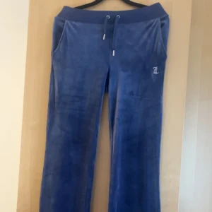 Blå velourbyxor från Juicy Couture - Blåa mjukisbyxor i velour från Juicy Couture med snörning i midjan och fickor. Klassisk Juicy-logga i strass på baksidan och broderad JC-logga fram. Sköna och snygga med loose passform och bred resår i midjan. Knappt Använda 