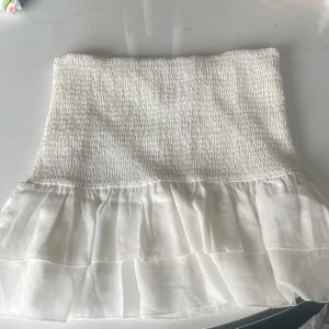 Vit volangkjol med smock - Supersöt vit kortkjol med smockad midja och dubbla volanger nertill. Kjolen är luftig och lätt, perfekt för varma dagar. Den har en romantisk vibe och passar till crop tops eller stickade tröjor  .DU BETALAR FRAKT