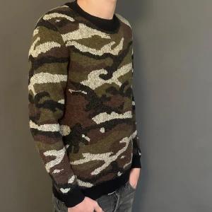 Saint Laurent stickad camouflage-tröja - Snygg stickad tröja från Saint Laurent med camouflage-mönster i brunt, grönt, svart och beige. Rund halsringning med ribbade muddar vid ärmslut och nederkant. Perfekt för dig som vill sticka ut med en exklusiv streetstyle-look.