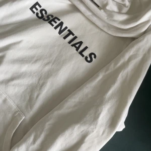 Vit Essentials hoodie från Fear of God - Säljer en stilren vit hoodie från Fear of God Essentials. Tröjan har svart tryck på bröstet och ärmen, klassisk huva och känguruficka. Mjuk bomullskvalitet och relaxed fit gör den perfekt för chill dagar. Essentials-logga även på ryggen.