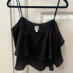 Svart volangig linnetopp från H&M - Supersnygg svart linnetopp från H&M med dubbla volanger och tunna justerbara axelband. Toppen har en luftig och flowy känsla, perfekt för varma dagar eller när du vill ha en chill men ändå stilren look.