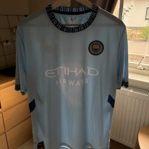 Manchester City Haaland 9 Puma tröja - Manchester Citys officiella ljusblå matchtröja med Haaland 9 på ryggen. Puma-logga på bröstet, klubbmärke och Etihad Airways-tryck framtill. Mörkblå detaljer på ärmslut och krage med unik design. Tillverkad i lätt och ventilerande material.