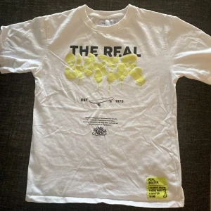 Vit t-shirt med skatertryck från Zara - Vit t-shirt från Zara med coolt skaterinspirerat tryck i svart och neongult framtill. Texten 'THE REAL SKATER' och skateboardgrafik ger streetkänsla. Rund halsringning och korta ärmar. Materialet är mjuk bomull, perfekt för en avslappnad look.