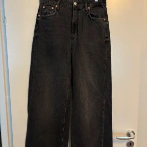Svarta wide jeans från Gina Tricot - Säljer ett par svarta jeans från Gina Tricot i modellen Perfect Jeans. De har hög midja, raka vida ben och klassisk femficksdesign. Jeansen är i ett mjukt denimtyg och stängs med dragkedja och knapp. Perfekta för en chill och trendig look.