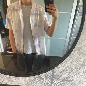 Ljuslila och vit skjorta i bomull - Kortärmad skjorta i bomull med ljuslila och vita inslag, loose fit och klassisk krage. Skjortan har knappar framtill och ett unikt, nästan batik-liknande mönster som ger en chill vibe. Perfekt för varma dagar och enkel att styla över en t-shirt.