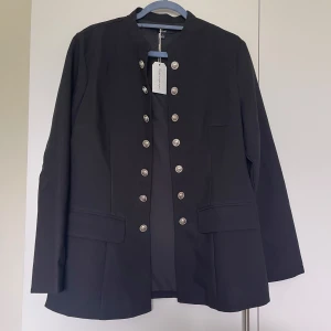 Svart rock med silverknappar - Stilren svart kavaj med raka linjer och dekorativa silverknappar framtill. Jackan har två falska fickor med lock och en kraglös design som ger en modern touch. Perfekt för dig som gillar en clean och snygg look.Helt ny med etikett 