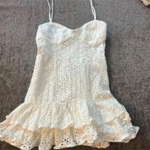 Vit spetsklänning med volang - Supersöt vit kortklänning med tunna axelband och volangkant nertill. Klänningen har broderad spets med hålmönster och en feminin, figurnära passform. Perfekt för sommarens alla tillfällen och ger en fräsch och romantisk look.