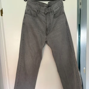 Grå jeans från G-Star RAW - Säljer ett par grå jeans från G-Star RAW i storlek (30/30) med raka ben och klassisk femficksdesign. Jeansen har hög midja och är tillverkade i mjukt denimtyg. Snygga detaljer med logga på bakfickan och metallknapp framtill. (Jeansen är ljusare i verkligheten)