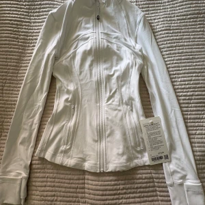Vit Define Jacket från Lululemon - Supersnygg vit Define Jacket från Lululemon med figurnära passform, hel dragkedja och långa ärmar med tumhål. Jackan är tillverkad i mjuk och stretchig polyesterblandning och har diskreta fickor med dragkedja. Perfekt för träning eller chill.