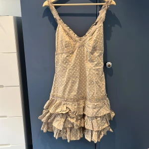 Beige prickig volangklänning - Supersöt kort klänning i beige med vita prickar och flera lager volanger nertill. Axelbandsmodell med v-ringning och rynkade detaljer. 
