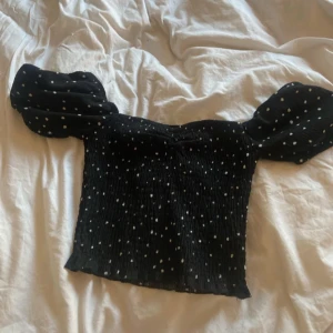 Svart prickig blus med puffärmar - Supersöt svart blus med vita prickar och stora puffärmar. Toppen har smockad kropp som sitter tajt och ger en snygg siluett. Perfekt för dig som gillar trendiga detaljer och vill sticka ut lite extra.