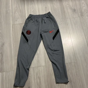 Grå PSG Nike träningsbyxor - Grå träningsbyxor från Nike med PSG-logga och röd swoosh på framsidan. Byxorna har svarta detaljer längs sidorna, elastisk midja och smal passform. Perfekta för fotboll eller chill, tillverkade i mjukt syntetmaterial.