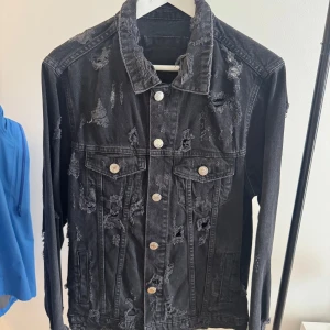 Svart  jeansjacka - Svart jeansjacka med slitna detaljer och trasiga partier för en edgy look. Klassisk modell med knappar framtill, två bröstfickor med lock och långa ärmar. Perfekt för dig som gillar streetstyle och vill sticka ut.