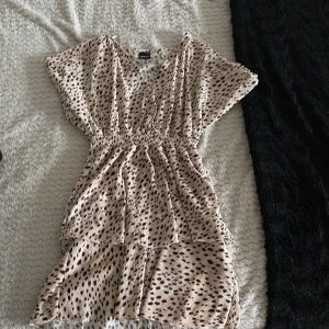 Beige kortklänning från Gina Tricot - Supersöt kortklänning från Gina Tricot i beige med svart prickigt mönster. Klänningen har v-ringning, resår i midjan och volangdetaljer nertill. Perfekt för sommarens alla tillfällen och har en luftig, skön passform.