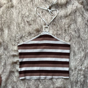 Randig ribbad halterneck topp Divided - Säljer en ribbad halterneck topp från Divided med breda bruna och vita ränder. Toppen har knyt i nacken och en fyrkantig ringdetalj framtill. Materialet är mjukt och stretchigt, perfekt för sommardagar. Snygg croppad passform som ger en trendig look.
