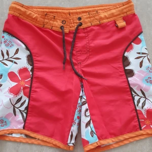 Badshorts - Snygga badshorts från Body Zone i rött och orange med blommigt mönster i vitt, rosa, blått och brunt. Shortsen har snörning i midjan och coola kontrastpaneler på sidorna. Perfekta för stranden eller poolen!