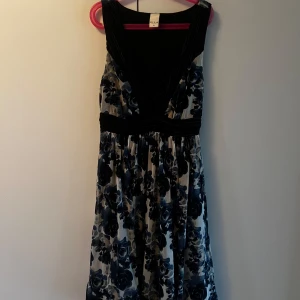 Blommig kortklänning från Vila XS - Supersöt kortklänning från Vila i XS med svart och blått blommönster. Djup v-ringning med snygga veckade detaljer och spetsinfällning upptill. Markerad midja och ärmlös design. Katt finns i hemmet. Kontakta vid fler frågor!🌷