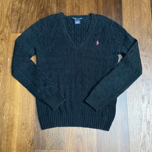 Svart kabelstickad tröja Ralph Lauren - Svart kabelstickad tröja från Ralph Lauren Sport med v-ringning och lång ärm. Tröjan har en rosa broderad logga på bröstet och är gjord i 100% bomull. Klassisk och stilren modell som är enkel att matcha. Kids size L. Längd 60 cm, bredd 42 cm. Fraktar oftast samma dag!