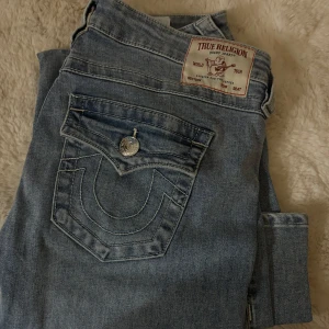 True Religion ljusblå bootcut jeans - Säljer ett par ljusblå jeans från True Religion med klassiska kontrastsömmar och stora fickor med lock och silverfärgade knappar. Jeansen har bootcut-modell och är tillverkade i mjukt denimtyg. Perfekta för dig som gillar street och en avslappnad vibe. Aldrig använda därav priset. Köpte en för liten storlek. 