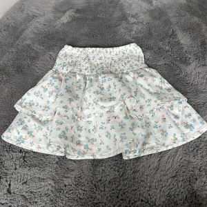 Blommig volangkjol från Gina Tricot - Supersöt kort kjol från Gina Tricot med blommigt mönster i pastellfärger på vit botten. Kjolen har bred smock i midjan och dubbla volanger som ger en flowy känsla. Perfekt för vår och sommar när du vill ha en lätt och luftig look.