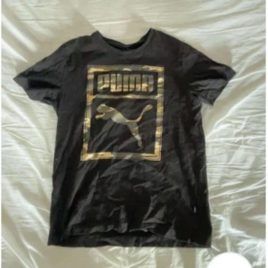 Svart Puma t-shirt med guldtryck - Svart t-shirt från Puma med stort guldigt logotryck framtill. Klassisk passform med rund hals och korta ärmar. Perfekt för dig som gillar sportiga och stilrena plagg med en touch av bling.